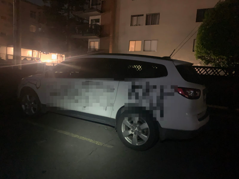 A white SUV is parked at night. The camera flash shows graffiti spray painted across the vehicle in black. In this photo the graffiti has been blurred out.
