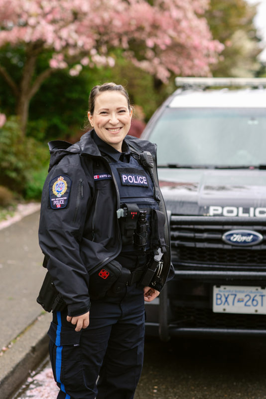 Sergeant Thom stands in uniform in front of an NWPD patrol car. She is smiling and is wearing a rain coat.