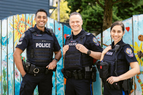 Three officers stand next to one another and smile. They are standing in front of a mural in the Uptown neighbourhood of New Westminster.