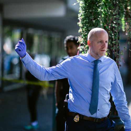 A Major Crime Unit detectives gives directives to other officers at a scene. He is described as male wearing a blue dress shirt and tie.