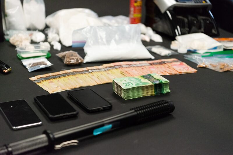 Numerous bags of drugs, weapons, a safe, and cash are displayed on a table. 