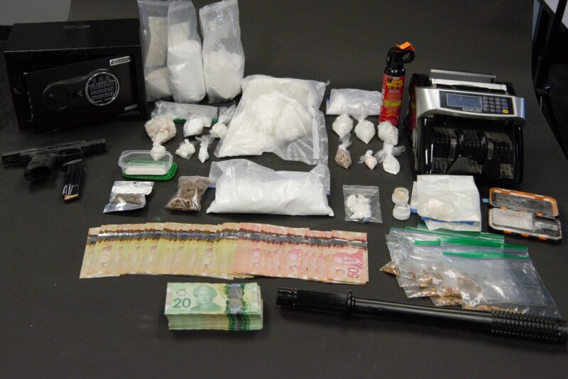 Numerous bags of drugs, weapons, a safe, and cash are displayed on a table. 